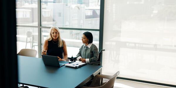 Two women, one blonde-haired and one with dark hair, discuss plans in a bright office space. The environment emphasizes professionalism, collaboration, and modern business interactions.
